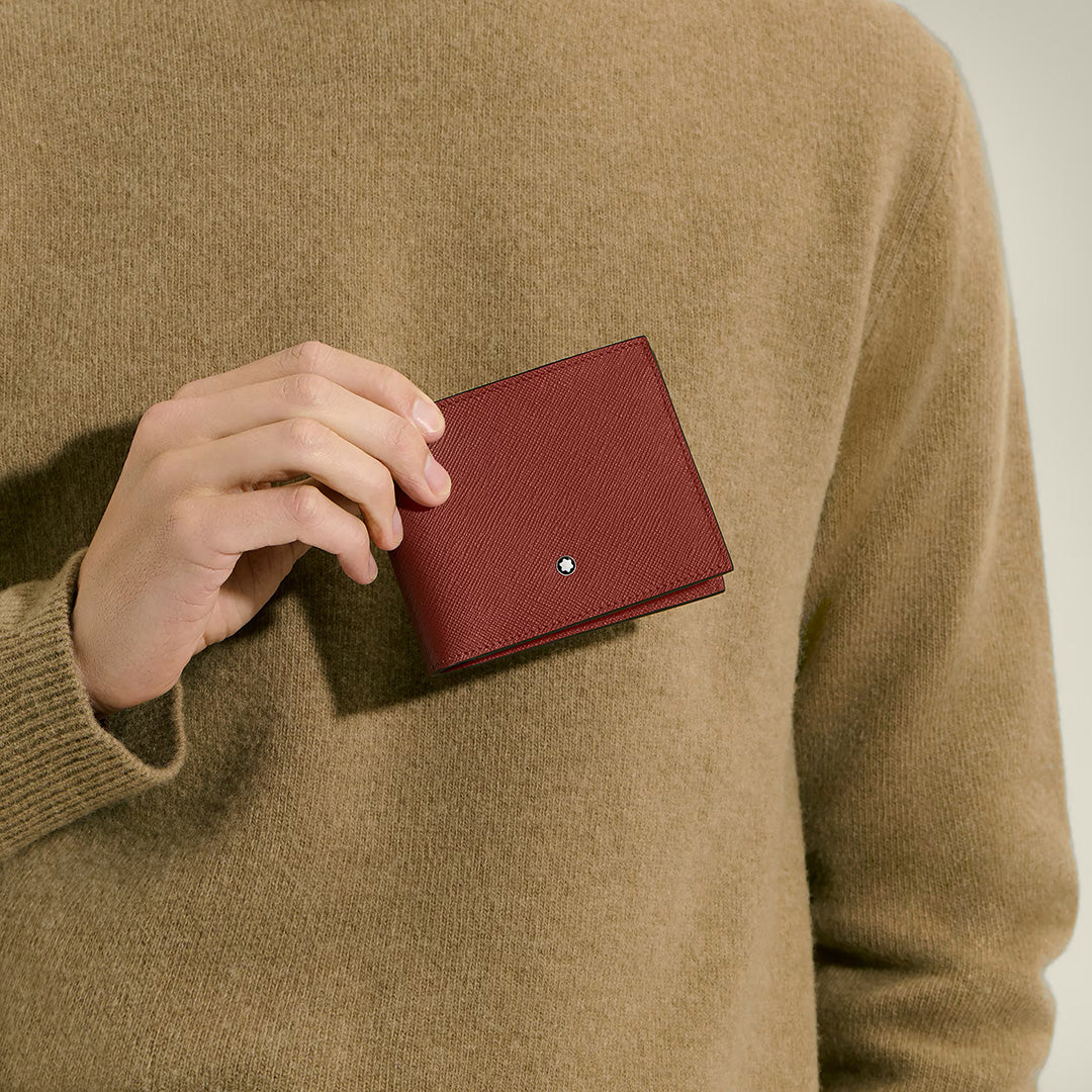 A person in a tan sweater holds the Montblanc Sartorial 6CC Leather Wallet—a closed red bifold featuring a small silver Montblanc emblem—near their chest, showcasing its refined embossed leather and signature Montblanc style.
