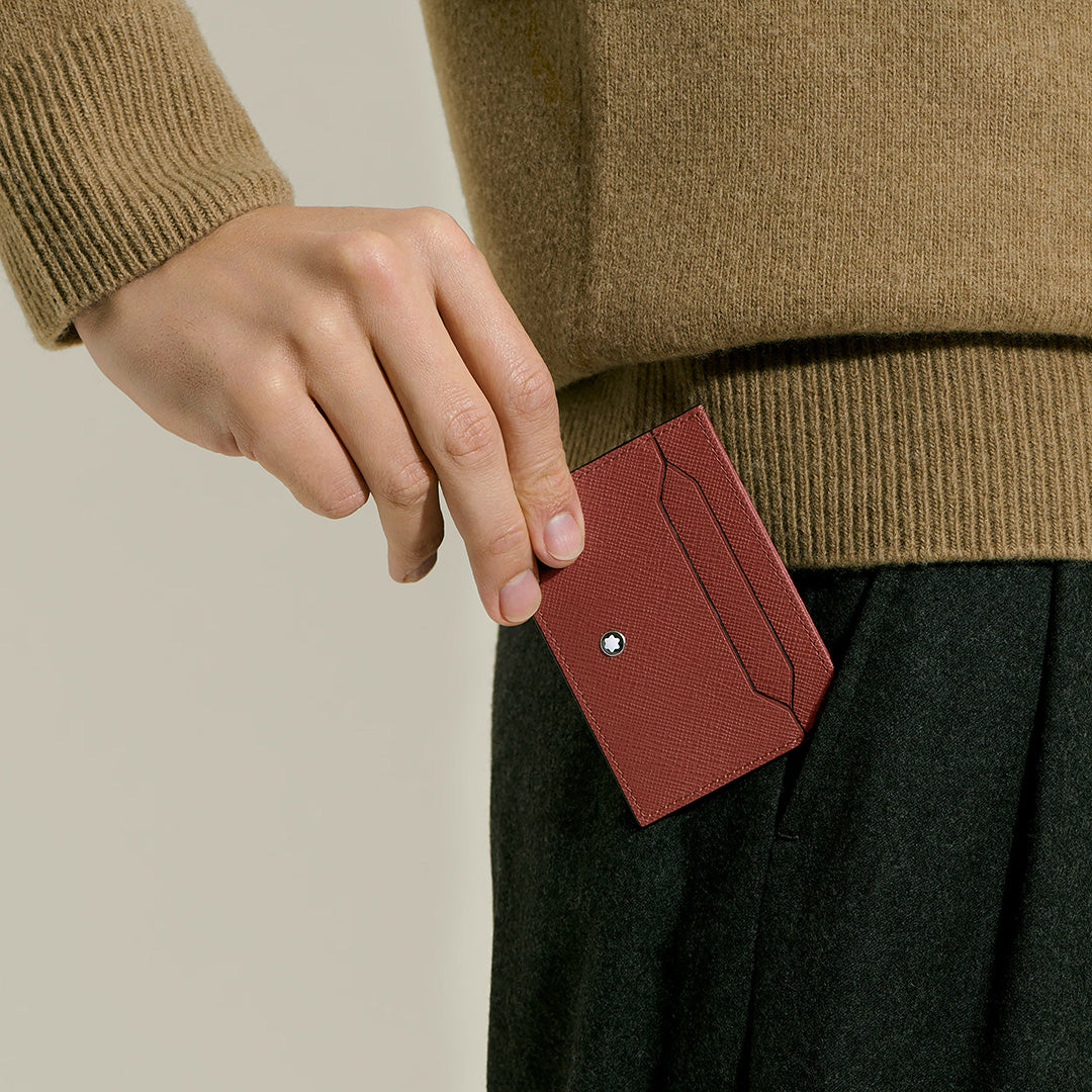 A person wearing a brown sweater and dark pants holds the Montblanc Sartorial 5CC Leather Card Holder, elegantly sliding the embossed Montblanc accessory into their pocket.