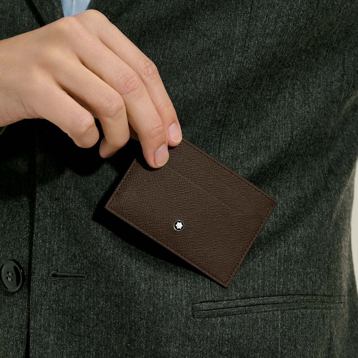 A person in a dark suit jacket slips a Montblanc Sartorial 5CC Leather Card Holder with an embossed finish and metal accent into their breast pocket.