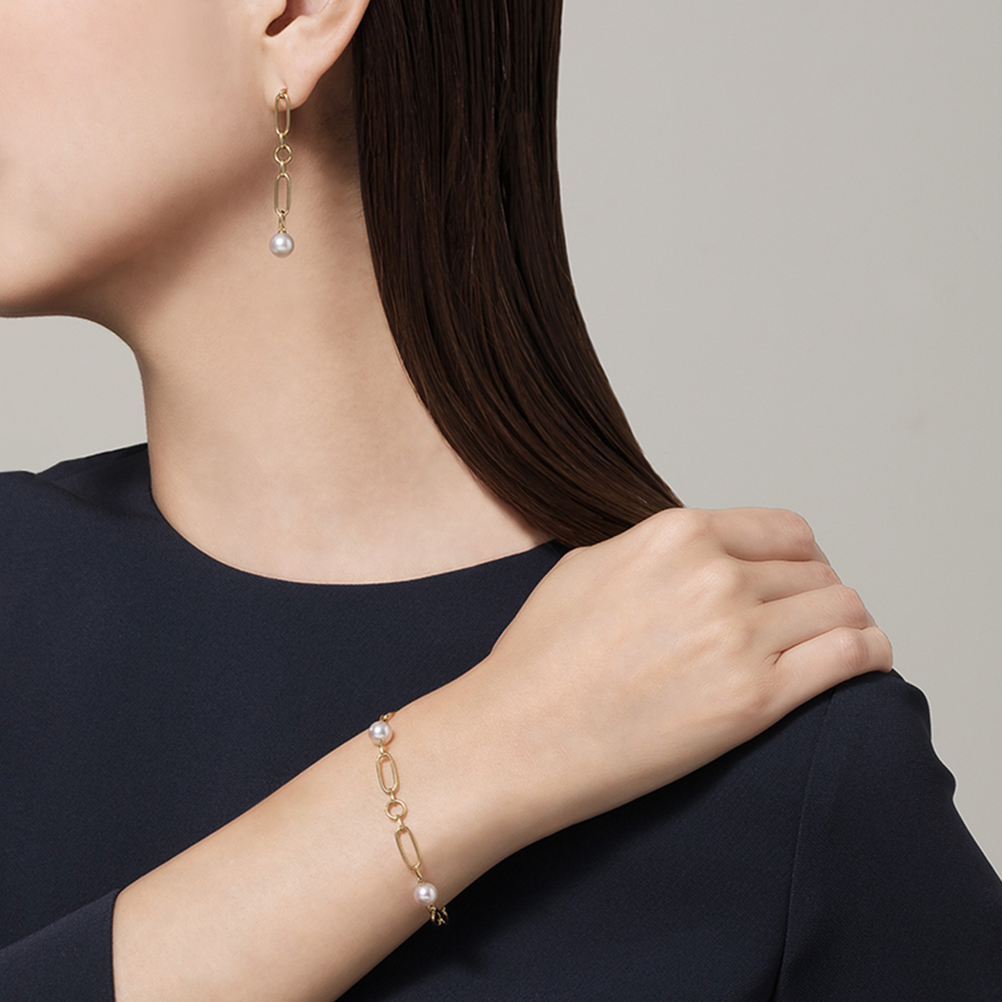 A person in a dark top displays elegant gold jewelry featuring Akoya cultured pearls, including a pair of dangling earrings and the Mikimoto M Code 18K Yellow Gold Akoya Cultured Pearl Station Bracelet. Their head is turned to the side, with their hand gently resting on their shoulder against a neutral background.