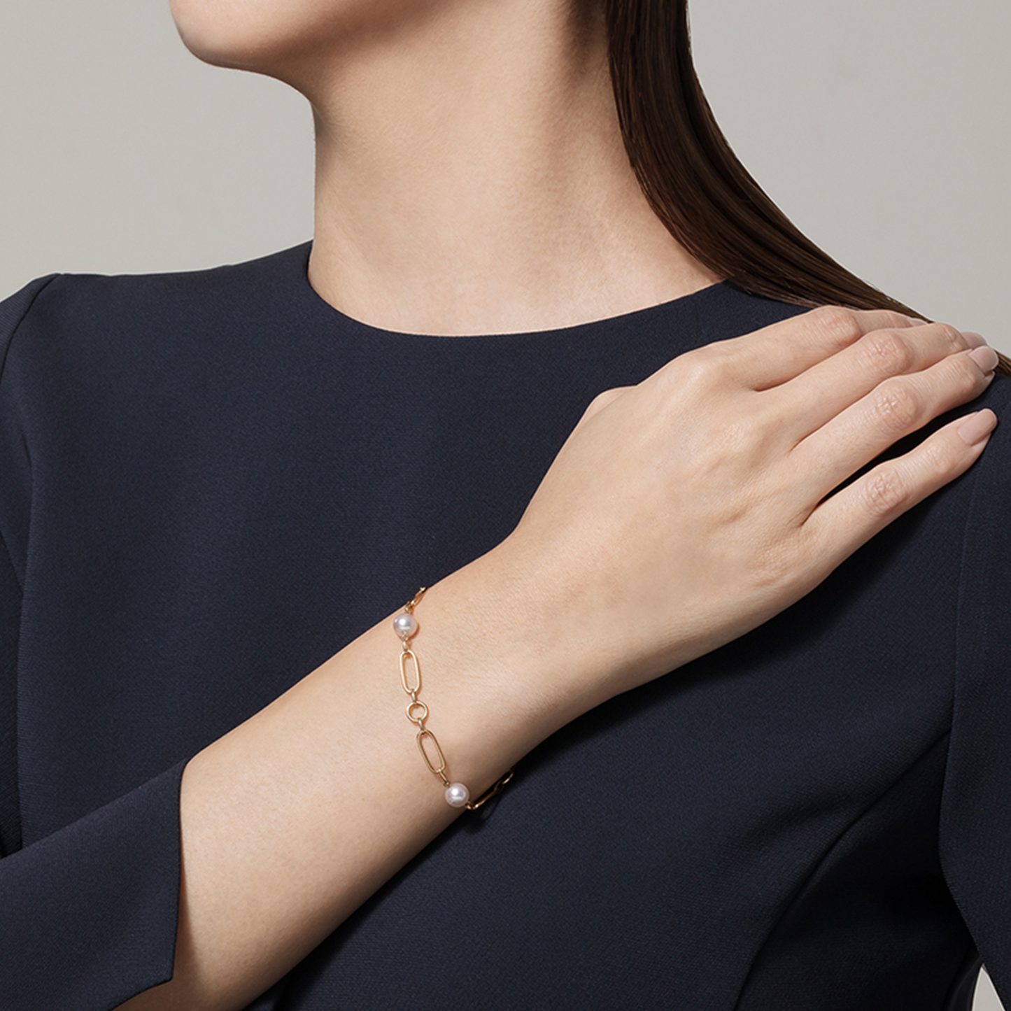 A woman in a dark dress, with her left hand resting on her right shoulder, displays the elegant Mikimoto M Code 18K Yellow Gold Akoya Cultured Pearl Station Bracelet. Her face is obscured, directing all focus to the stunning piece from Mikimoto's collection.