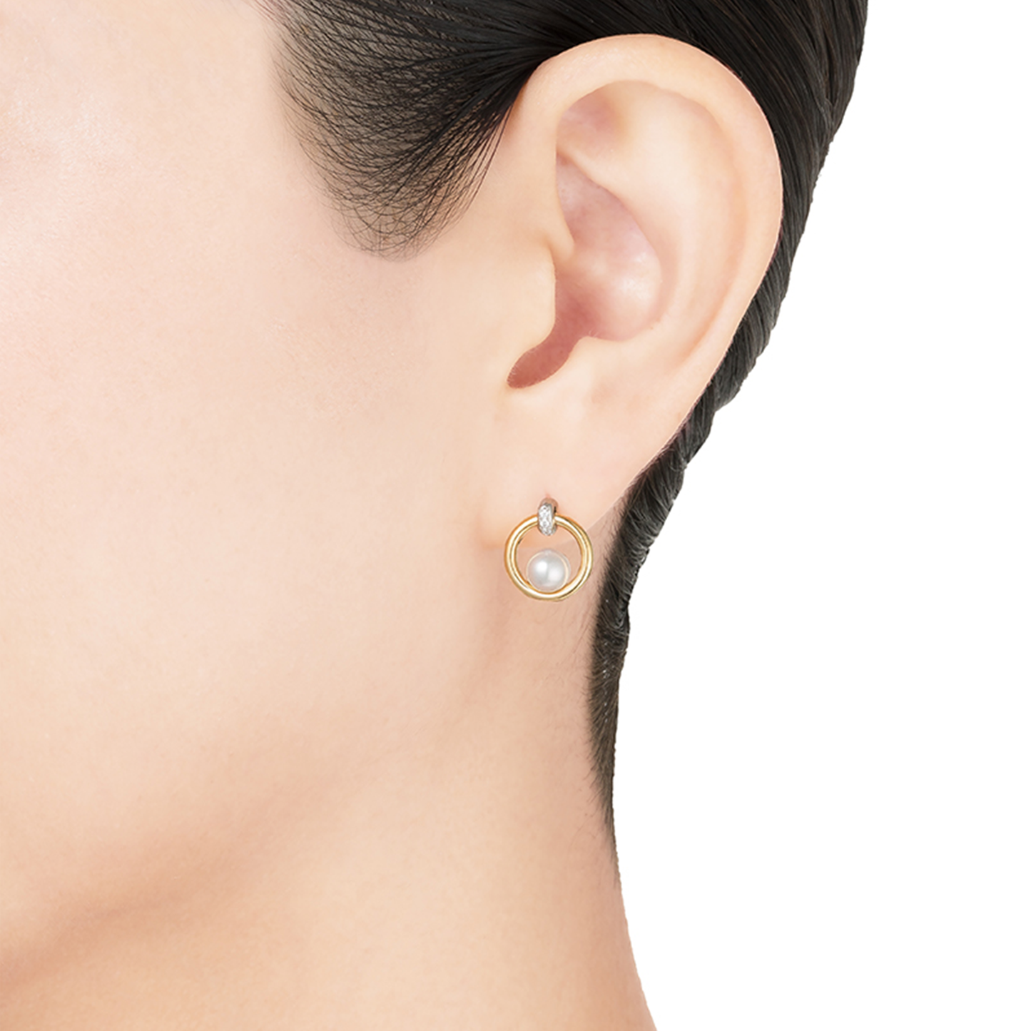 Close-up of a person's ear adorned with an elegant Mikimoto 18K Yellow and White Gold Diamond Akoya Cultured Pearl Circle Earring. The earring features a stunning Akoya pearl centered in a gold circular frame, highlighted by a small diamond at the top. The plain white background accentuates the refined design of this Mikimoto piece.