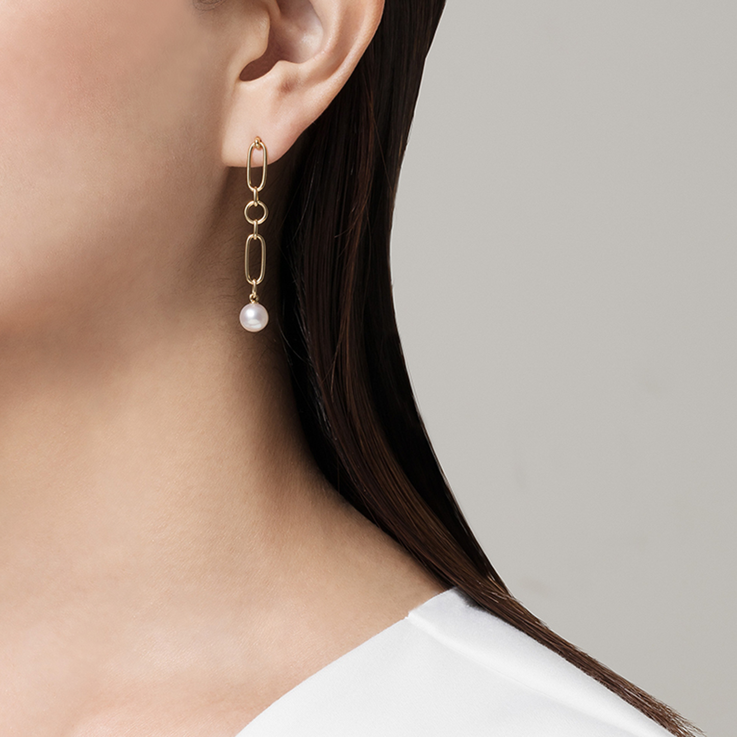 A close-up captures the contemporary allure of a person wearing the Mikimoto M Code 18K Yellow Gold Akoya Cultured Pearl Drop Earrings. The earring features a delicate chain and an Akoya cultured pearl, beautifully contrasting against their dark hair and white blouse, all set against a plain gray background.