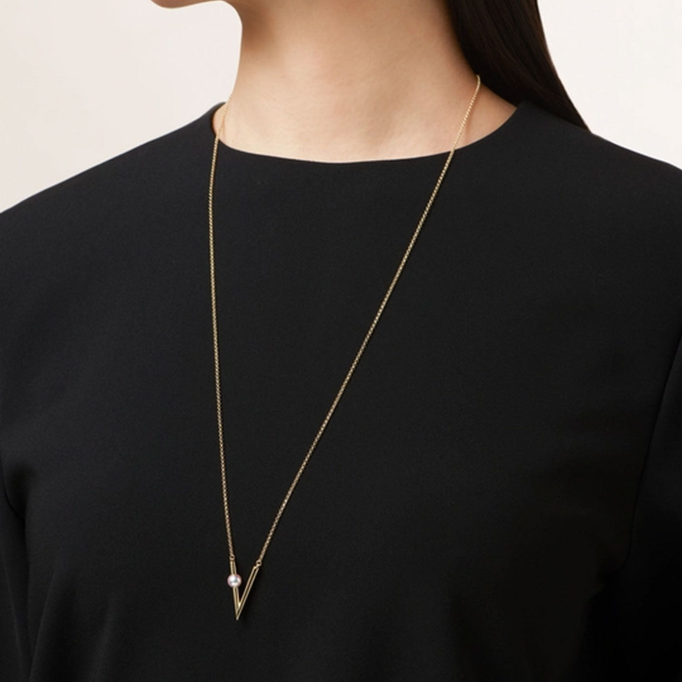 A person wearing a black top is shown with the Mikimoto V Code 18K Yellow Gold Akoya Cultured Pearl Necklace Pendant, which features a V-shaped pendant on an adjustable chain.