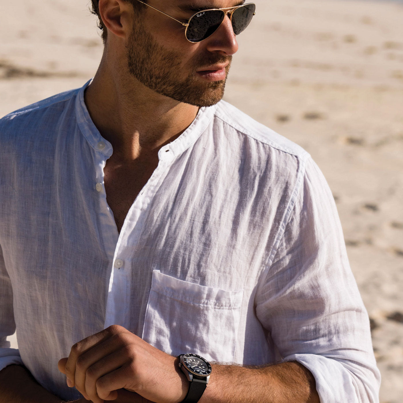 A person wearing sunglasses and a white button-up shirt stands on a sandy beach, checking their CITIZEN Promaster Fujitsubo Super Titanium Automatic Mechanical 41mm Watch. They glance to the side, with the bright, sunlit shoreline in the background.