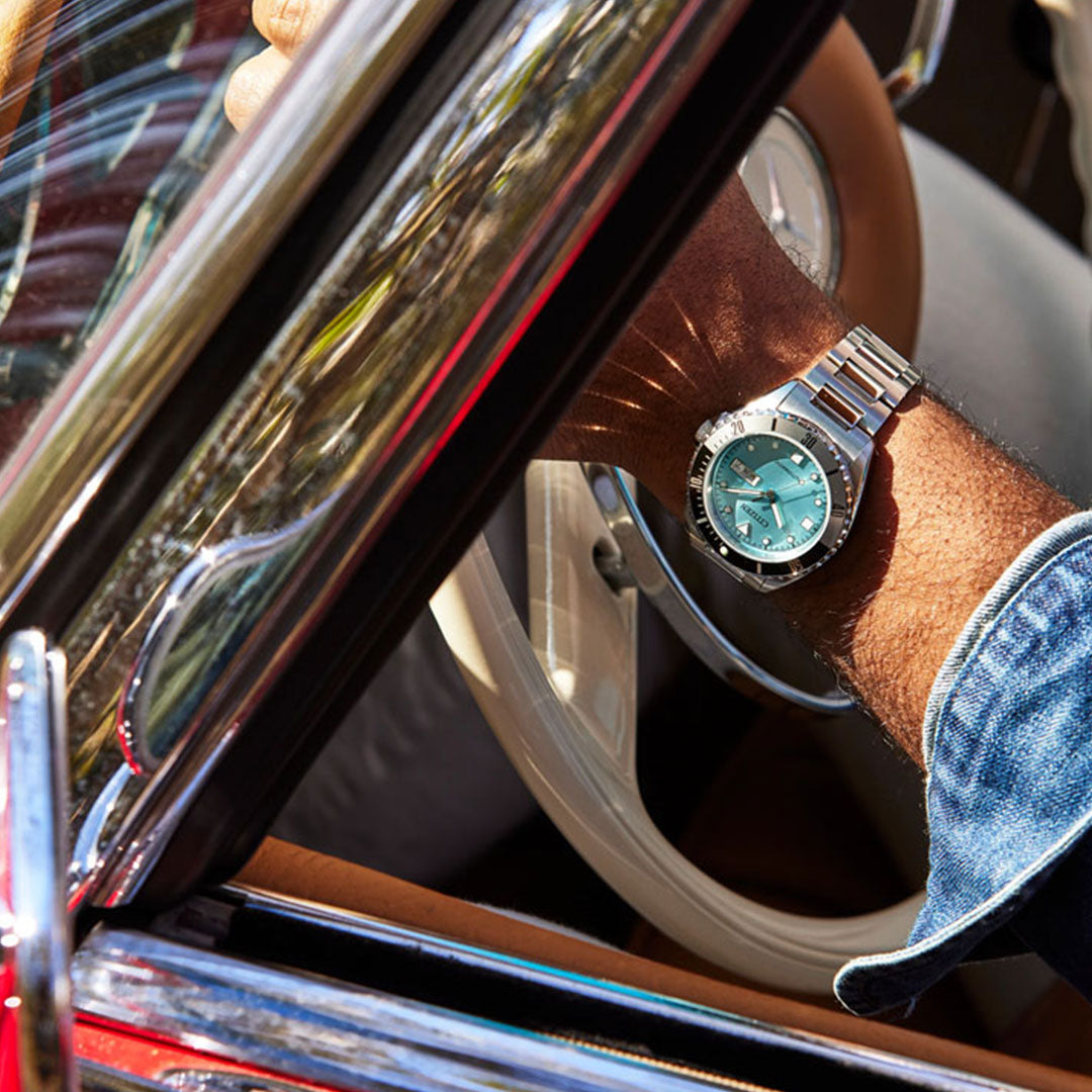 A person wearing the CITIZEN Sport Automatic 42mm Watch, which features a turquoise dial and sapphire crystal, steers a vintage car. The view captures the arm resting on the light-colored steering wheel, highlighting both the watch and the car's red exterior.