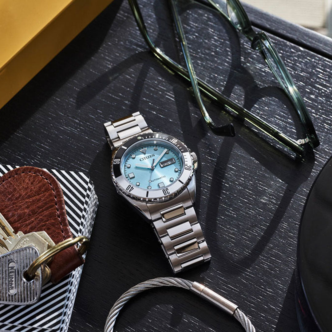 The CITIZEN Sport Automatic 42mm Watch, featuring a silver case, light blue face, and sapphire crystal, rests on a dark wooden surface alongside a pair of glasses, a leather wallet, keys, and a metal bracelet.