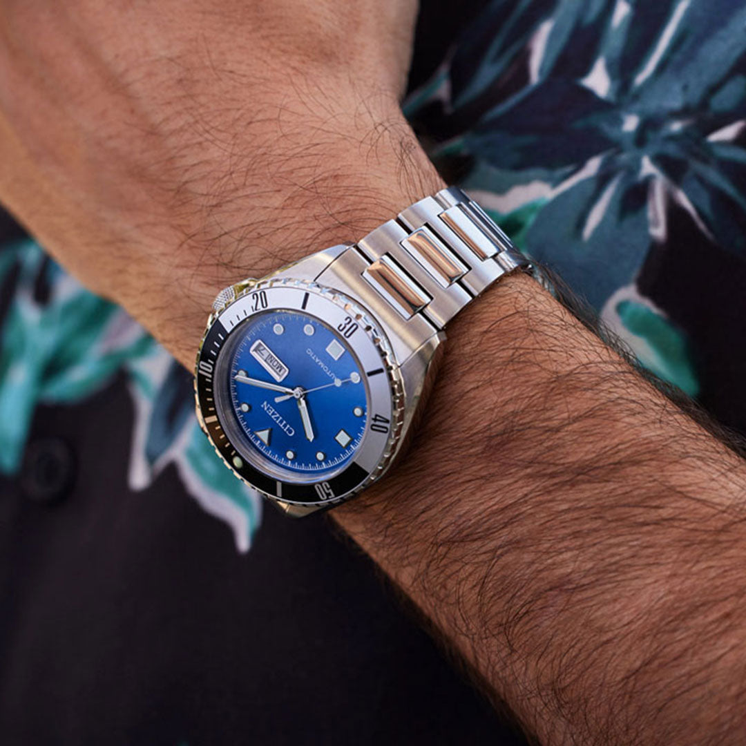 Close-up of a person's wrist wearing a CITIZEN Sport Automatic 42mm Watch in stainless steel featuring a blue dial and sapphire crystal. The watch showcases the day and date. The person is dressed in a dark, floral-patterned shirt.