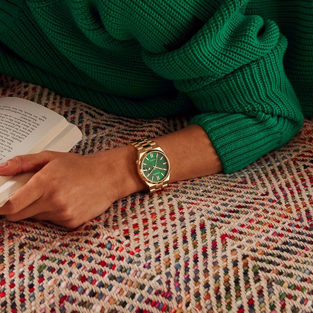 A person in a green sweater relaxes on a multicolored blanket with an open book, wearing a CITIZEN TSUYOSA Collection Automatic Mechanical 40mm Watch. The sunray green dial complements their sweater, and the gold-tone stainless steel band adds elegance to their ensemble.