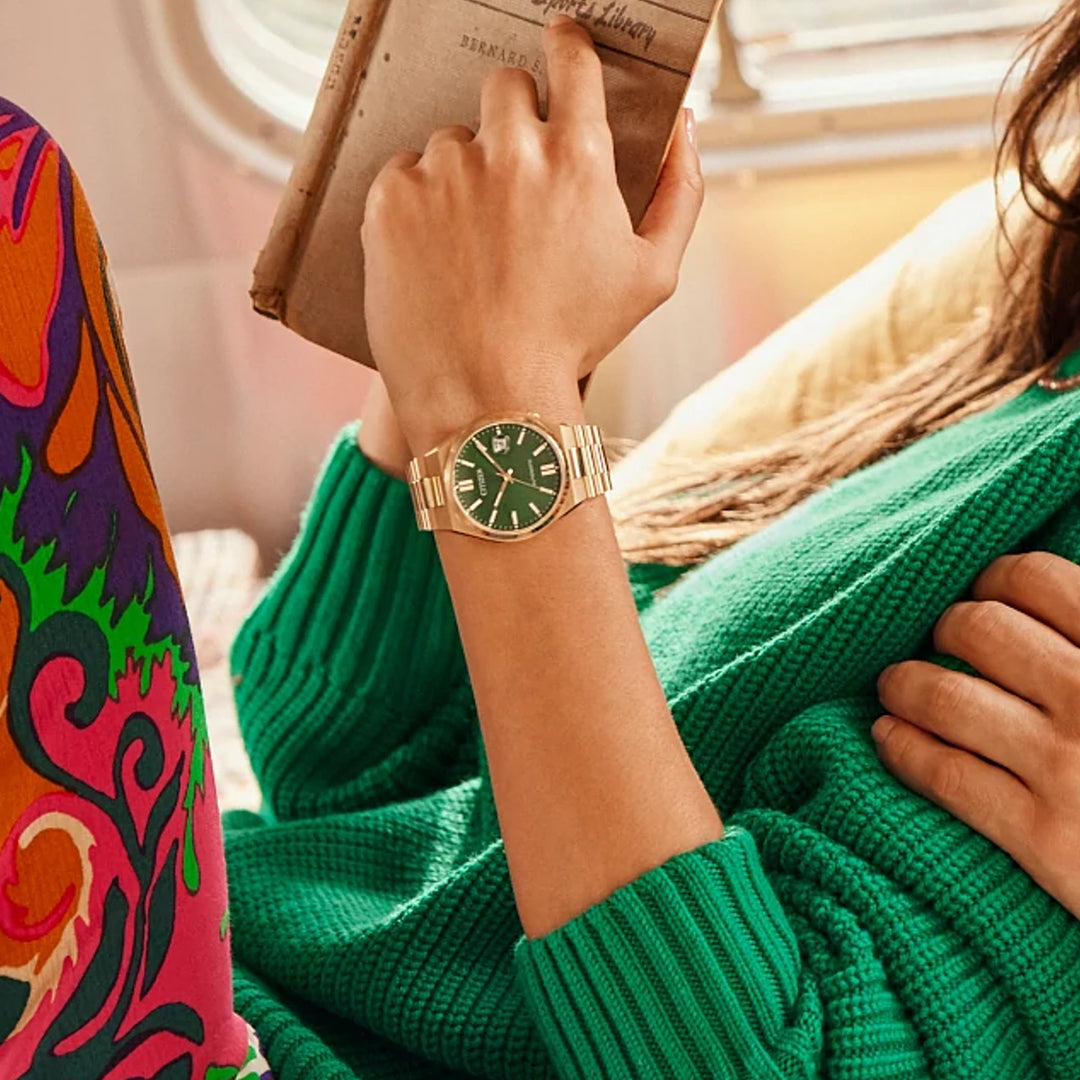 A person in a green sweater wears a CITIZEN TSUYOSA Collection Automatic Mechanical 40mm Watch with a sunray green dial and gold-tone stainless steel case, holding an open book while seated on a colorful patterned cushion.