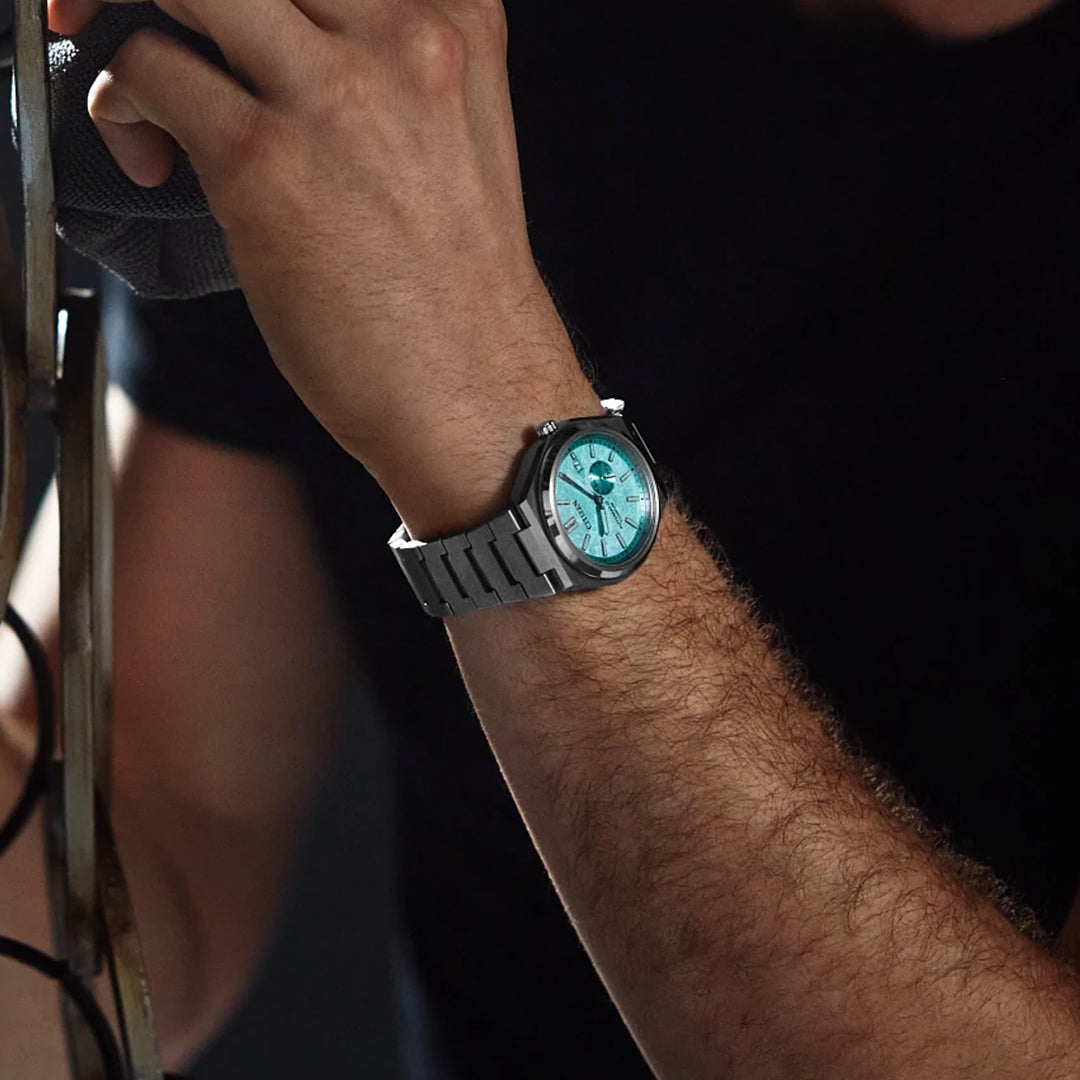 A person in a short-sleeved black shirt holds an object while wearing the CITIZEN Zenshin Mechanical Automatic 40.5mm Watch, a sporty stainless steel timepiece with a light blue dial and silver hour markers from CITIZEN.