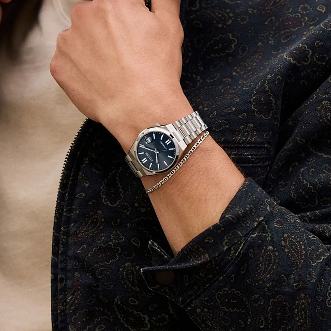 Wearing a dark patterned jacket and beige shirt, a person sports the CITIZEN TSUYOSA Collection Automatic 37mm Watch with sapphire crystal and silver bracelet, featuring a dark blue face, held near their chin.