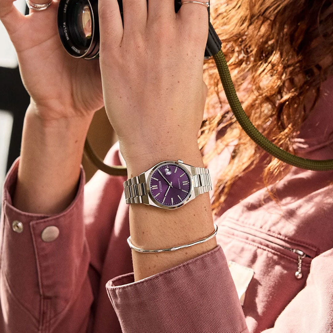 A person with curly hair in a mauve jacket holds a camera. On their left wrist, they wear a silver bracelet and the CITIZEN TSUYOSA Collection Automatic 37mm Watch, showcasing CITIZEN's elegant automatic design with its distinctive purple face.