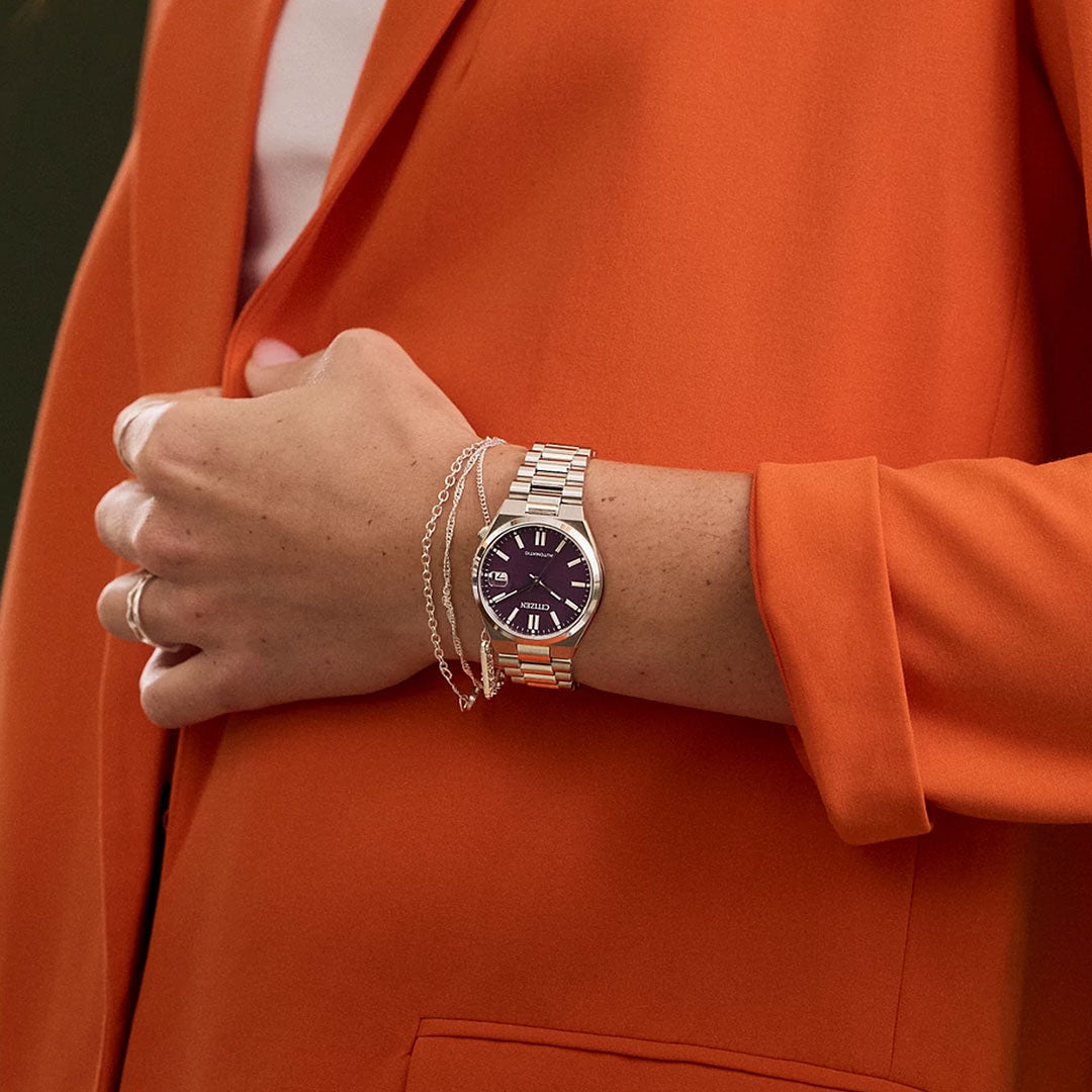 A person in an orange blazer wears a CITIZEN TSUYOSA Collection Automatic 37mm Watch with a dark face and silver band, paired with a delicate silver chain bracelet, shown with their arm bent and hand near their chest.