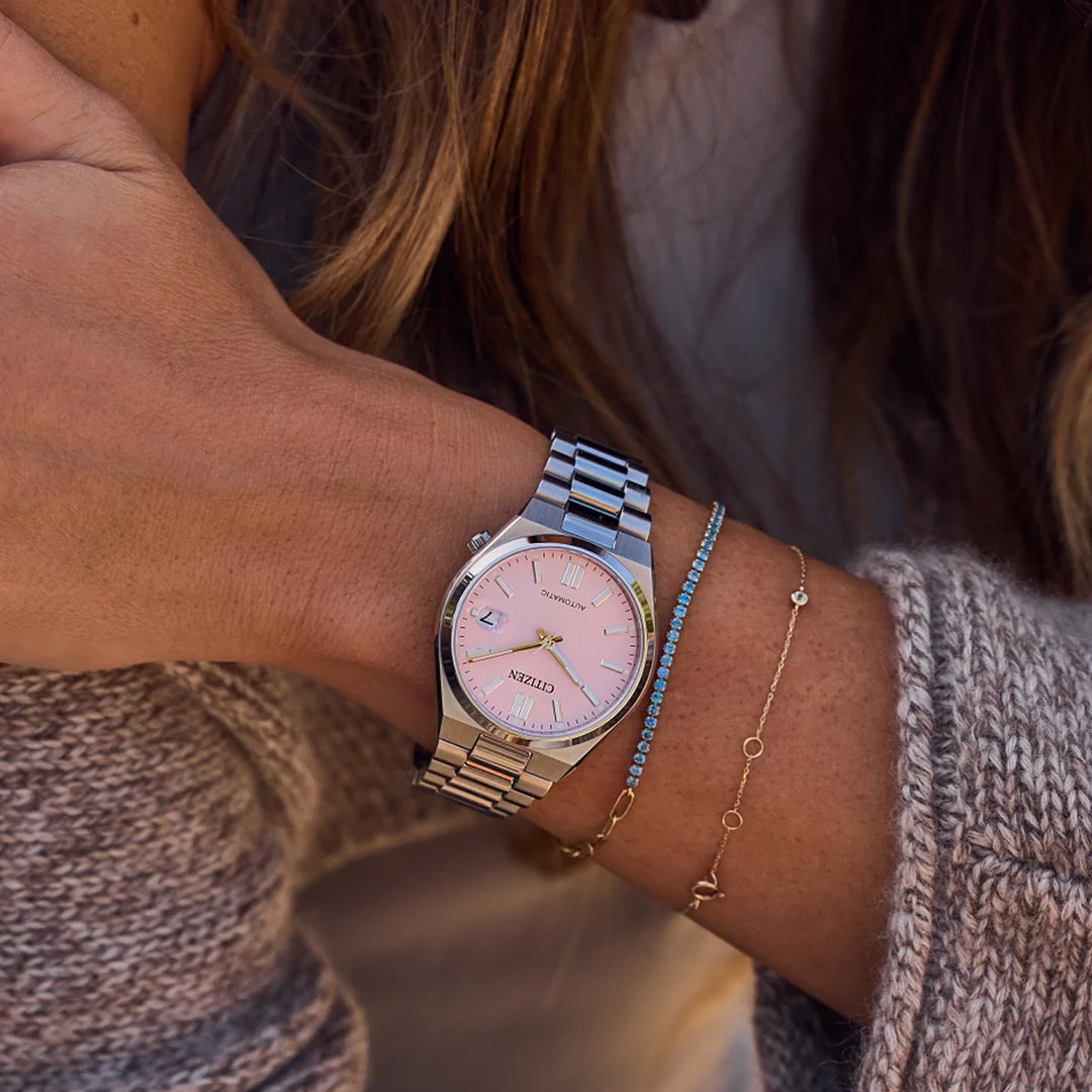 A person with long brown hair, dressed in a cream sweater, wears the CITIZEN TSUYOSA Collection Automatic 37mm Watch with a pastel pink dial and stainless steel case, paired with two delicate gold bracelets.