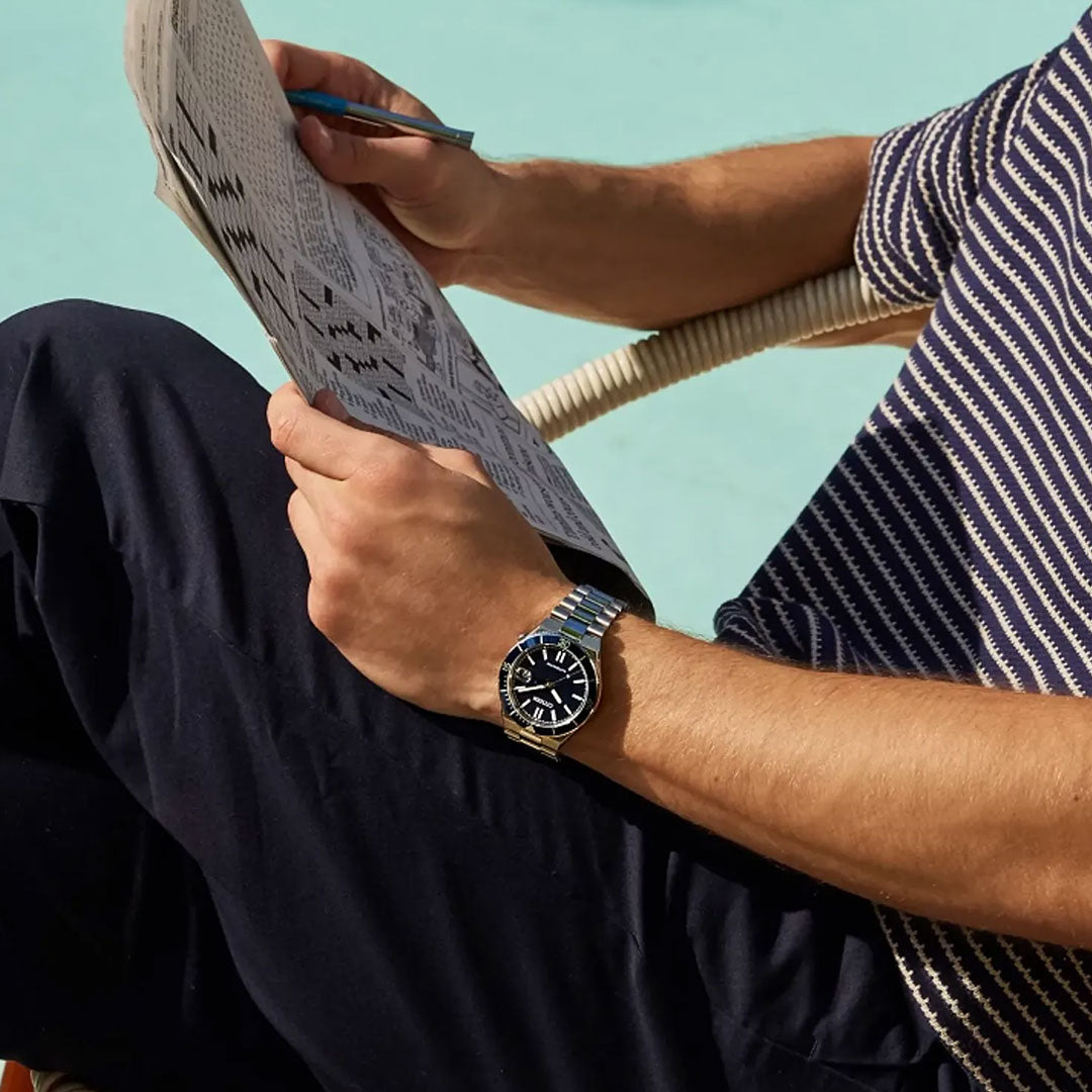 A person sits by the pool, working on a crossword puzzle in a striped short-sleeve shirt, dark pants, and wearing the CITIZEN Tsuyosa Shore Automatic 40mm Watch from CITIZEN.