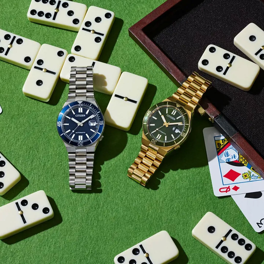 Two CITIZEN wristwatches, including the CITIZEN TSUYOSA Shore Automatic 40mm with a gold-tone stainless steel case and black dial, rest on a green surface among dominoes and partly visible playing cards.