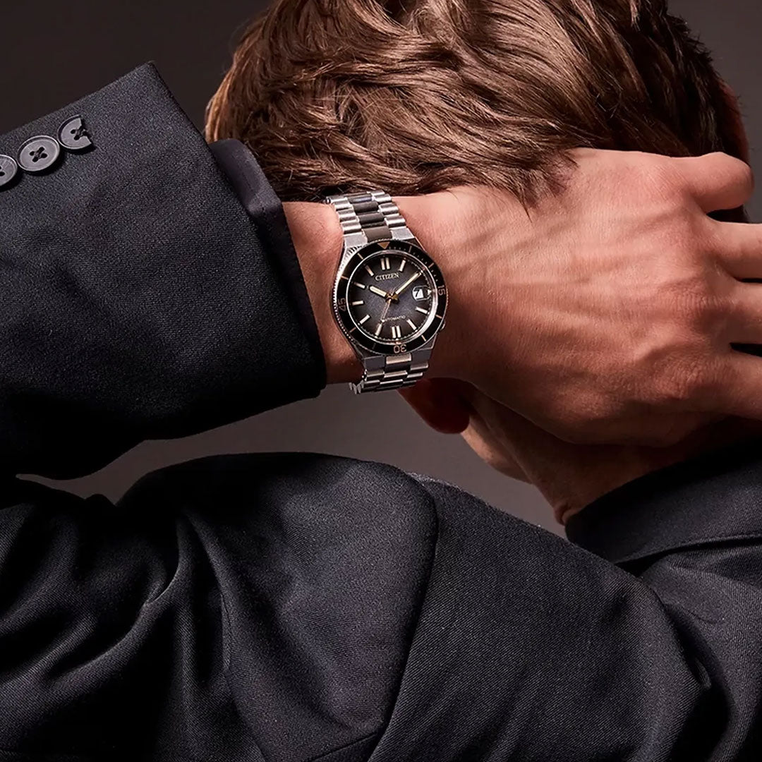 A person in a black suit jacket poses with their hand on the back of their head, showcasing a CITIZEN TSUYOSA Shore Automatic 40mm Watch “Time Slip” featuring a silver wristband, black face, gold hour markers, and an automatic calibre 8210.