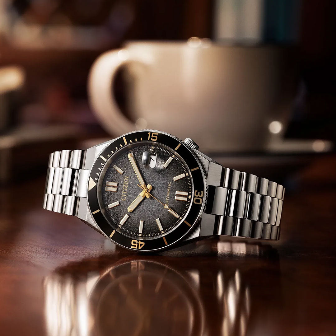 A CITIZEN TSUYOSA Shore Automatic 40mm Watch “Time Slip” by CITIZEN, featuring a silver metal band and black dial with the precise automatic calibre 8210 movement, rests on a polished wooden surface; in the background, a blurred coffee cup and saucer complete the scene—an understated yet modern sports watch suitable for any occasion.