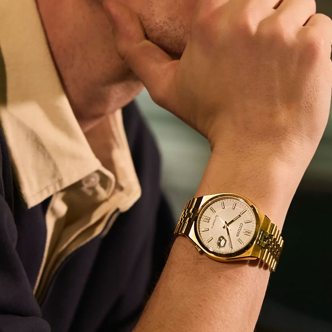 A person wearing the CITIZEN TSUYOSA 60 Automatic 40mm Watch by CITIZEN, featuring a yellow gold-tone case and a white dial, rests their chin on their hand while dressed in a collared shirt and dark sweater.