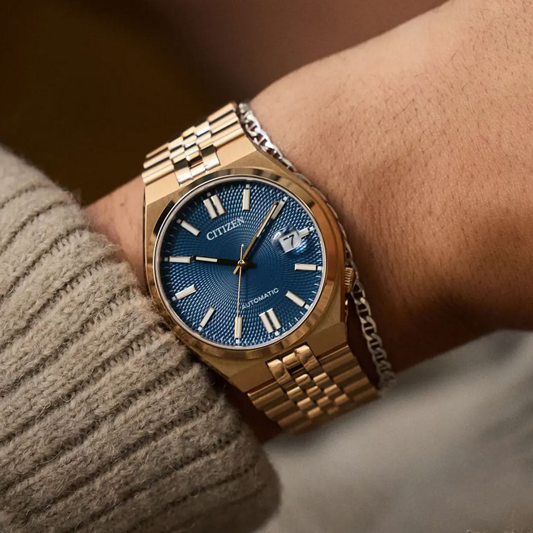 A CITIZEN TSUYOSA 60 Automatic 40mm gold watch with a blue textured dial and date display is worn beside a silver chain bracelet. The wearer has on a beige ribbed sweater.