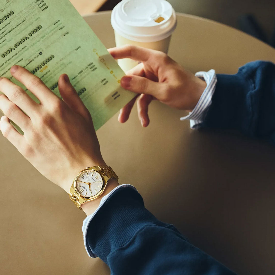 A person wearing the CITIZEN TSUYOSA 60 Automatic 37mm Watch by CITIZEN, which features a yellow gold-tone case and an automatic calibre 8310, holds a green menu at a round table with a takeaway coffee cup, dressed in a dark sweater and striped shirt.