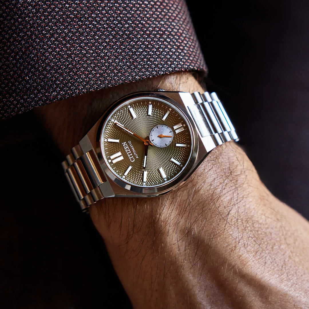 A person wears the CITIZEN TSUYOSA Small Second Automatic 40mm Watch with a stainless steel case, textured gold dial, silver markers, and a small seconds subdial on their left wrist against a brown patterned shirt sleeve.