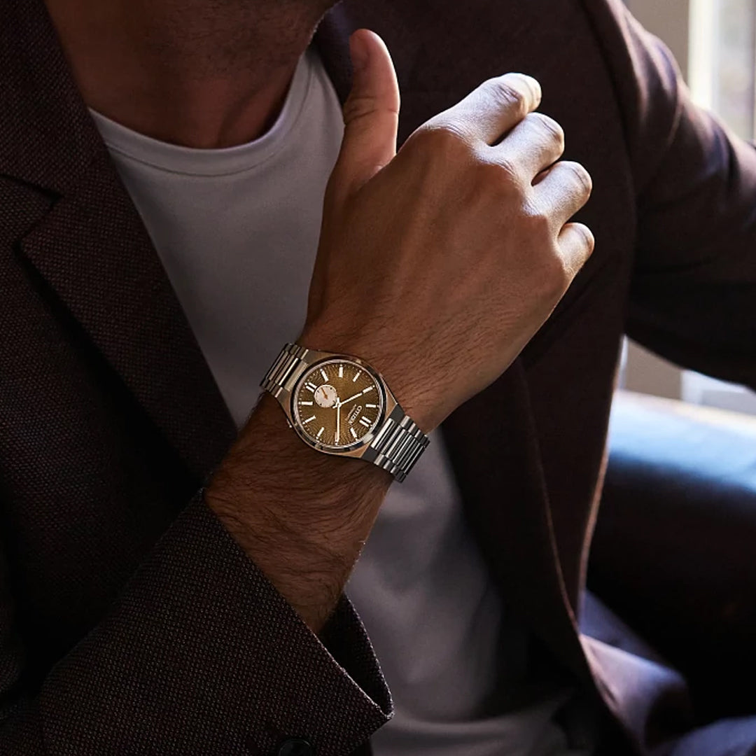 A person in a brown blazer and white shirt displays the CITIZEN TSUYOSA Small Second Automatic 40mm Watch with a green dial and stainless steel case, relaxing with an arm on a chair.