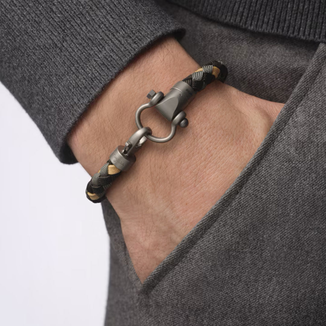 A person in a dark gray sweater has their hand in their pocket, revealing an OMEGA Aqua Sailing 007 Edition Nylon Bracelet with black and beige braiding and a metallic clasp on their wrist.