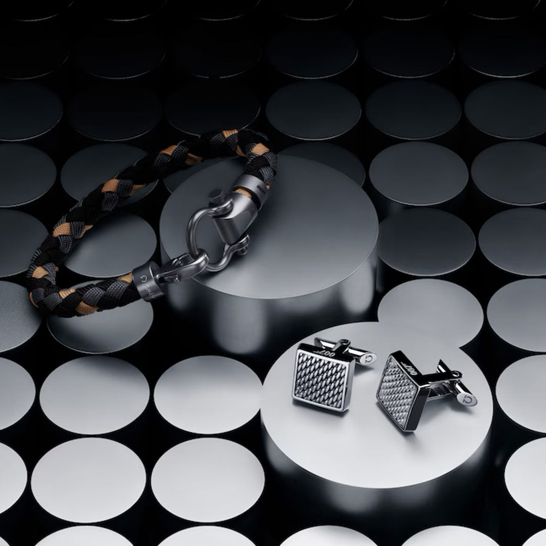 The OMEGA Aqua Sailing 007 Edition Nylon Bracelet rests on a round, reflective silver platform, paired with a set of square patterned cufflinks, against cylindrical silver shapes.
