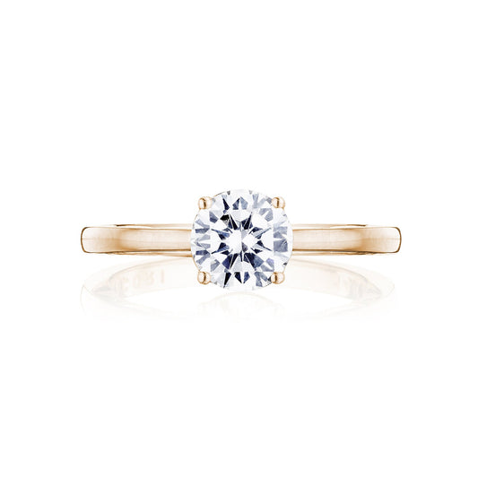 A 14K rose gold engagement ring from the Tacori Coastal Crescent collection is displayed, featuring a round brilliant solitaire diamond. Crafted with simplicity and elegance, the band prominently showcases the diamond at its center with delicate accents. The image is set against a pristine white background.