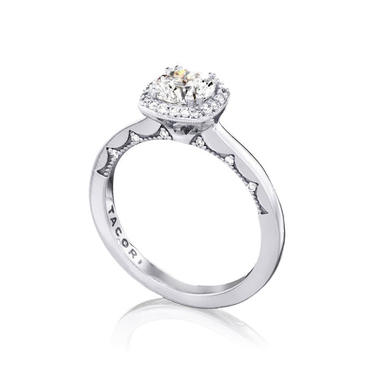The Tacori Coastal Crescent engagement ring in 14K white gold features a central large round brilliant diamond, surrounded by a halo of smaller diamonds. The band is intricately detailed and engraved with "TACORI," embodying elegance and timeless beauty.