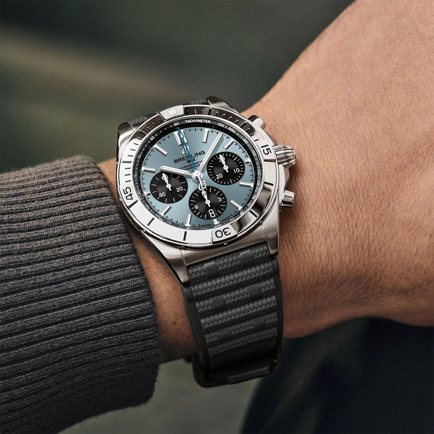 A close-up of a person's wrist wearing the luxurious BREITLING Chronomat B01 42 by Breitling, showcasing its silver case and blue dial. This exquisite watch features three subdials and a black strap, perfectly complementing their gray, ribbed long-sleeve shirt. The softly blurred backdrop enhances the elegance of this timepiece.