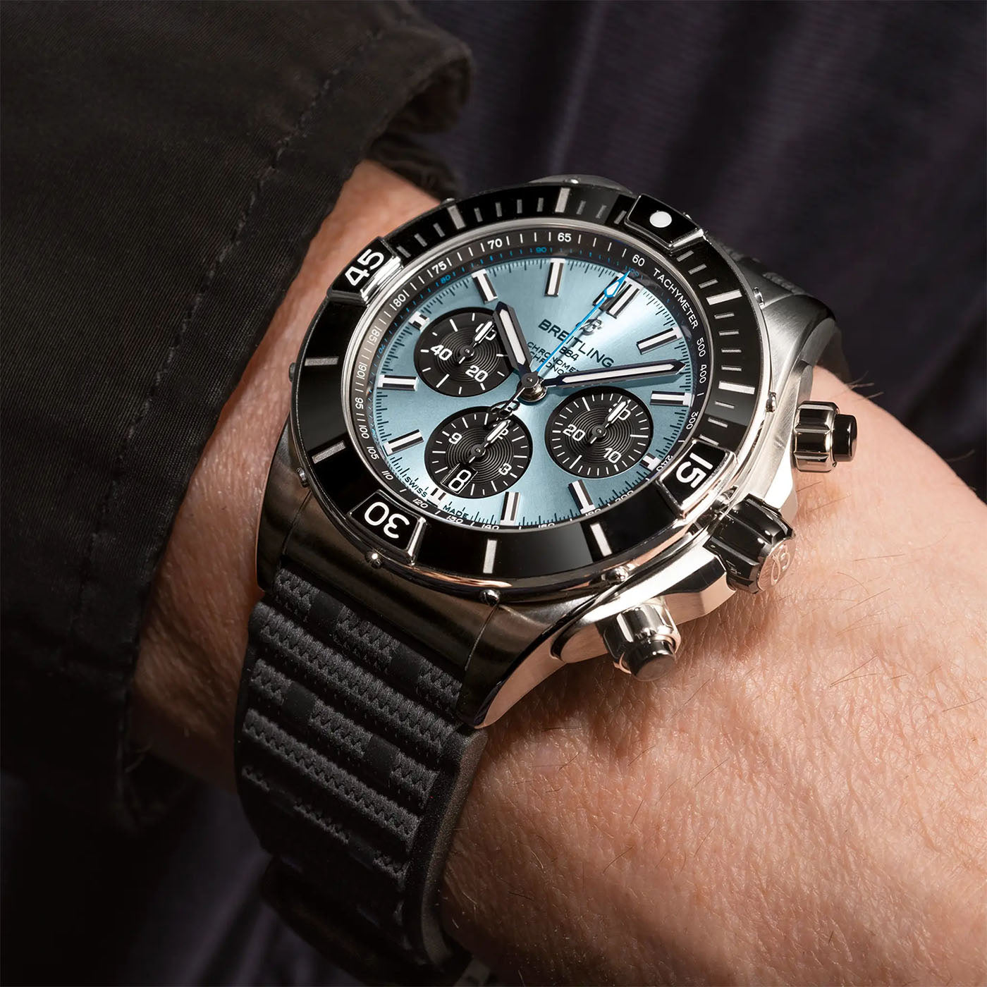 The BREITLING Super Chronomat B01 44 wristwatch features a stainless steel case, black bezel, and light blue dial with three subdials. It is adorned with lab-grown diamonds and has a black textured strap. A dark sleeve complements the wearer's arm in the close-up shot.