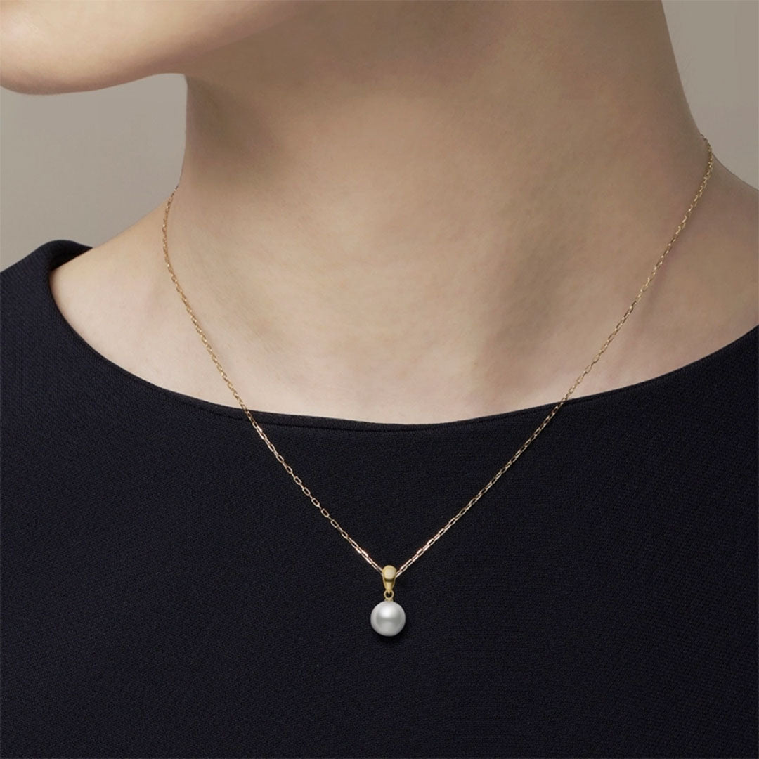 A close-up of a person wearing the Mikimoto 18K Yellow Gold Akoya Cultured Pearl Necklace Pendant, featuring a single round 6-6.5mm Akoya cultured pearl, shown against a plain dark top.