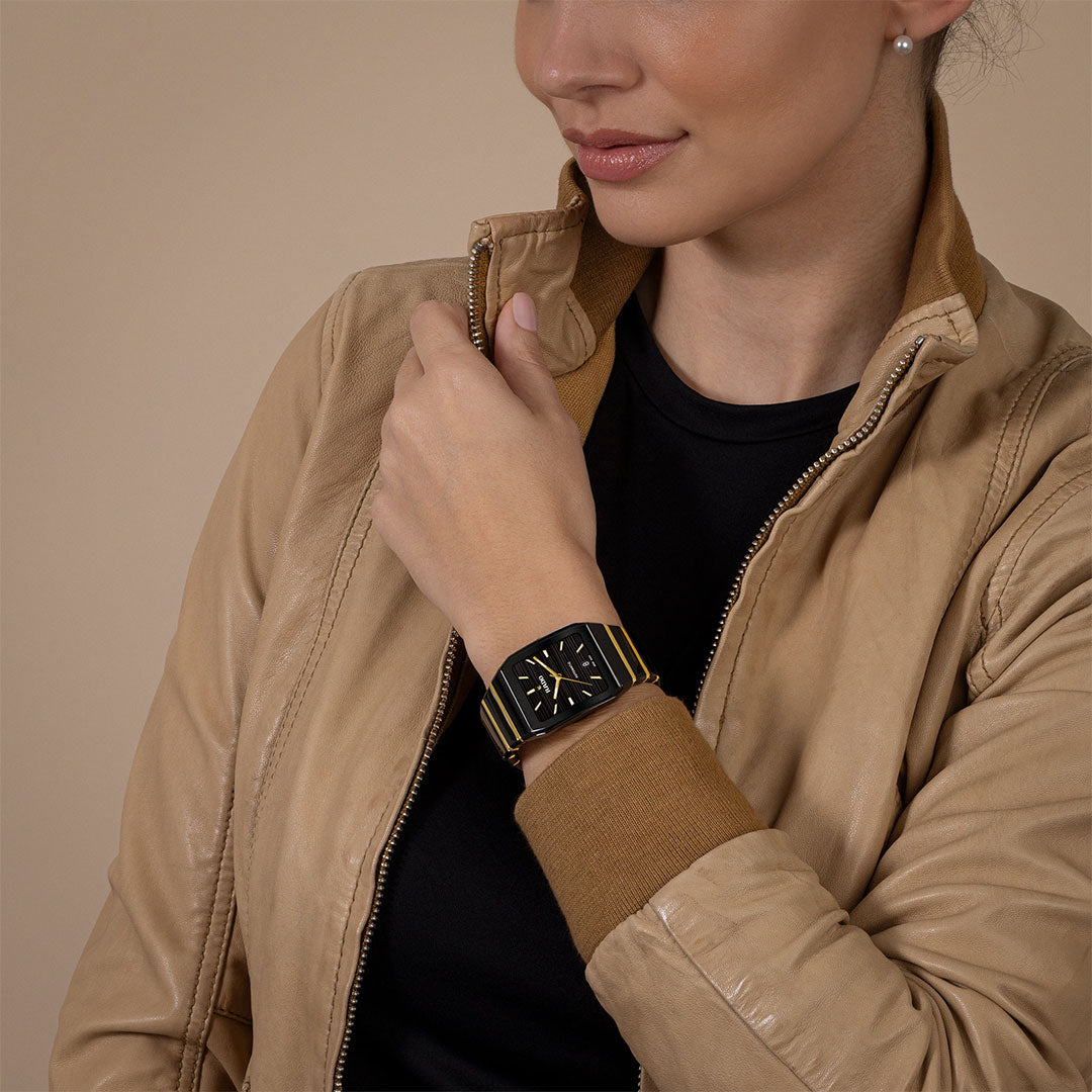 A woman in a tan leather jacket and black shirt displays the RADO Anatom Automatic 32.5mm Watch, featuring a high-tech ceramic bracelet and sapphire crystal, its black face with yellow stripes contrasting against a neutral backdrop.