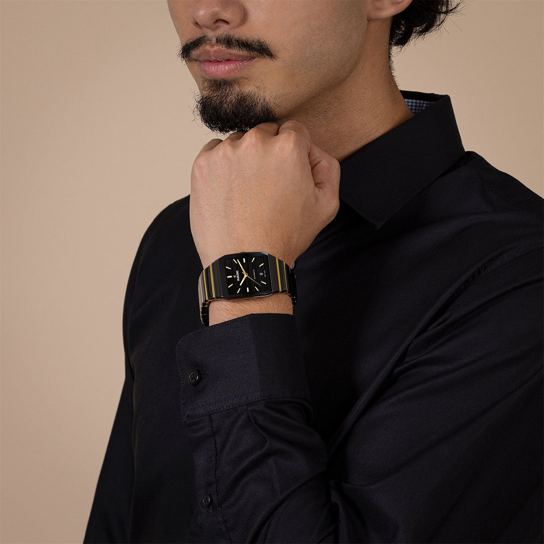 A man in a black dress shirt poses with his hand near his face, wearing the RADO Anatom Automatic 32.5mm watch by RADO on his left wrist against a plain beige background.