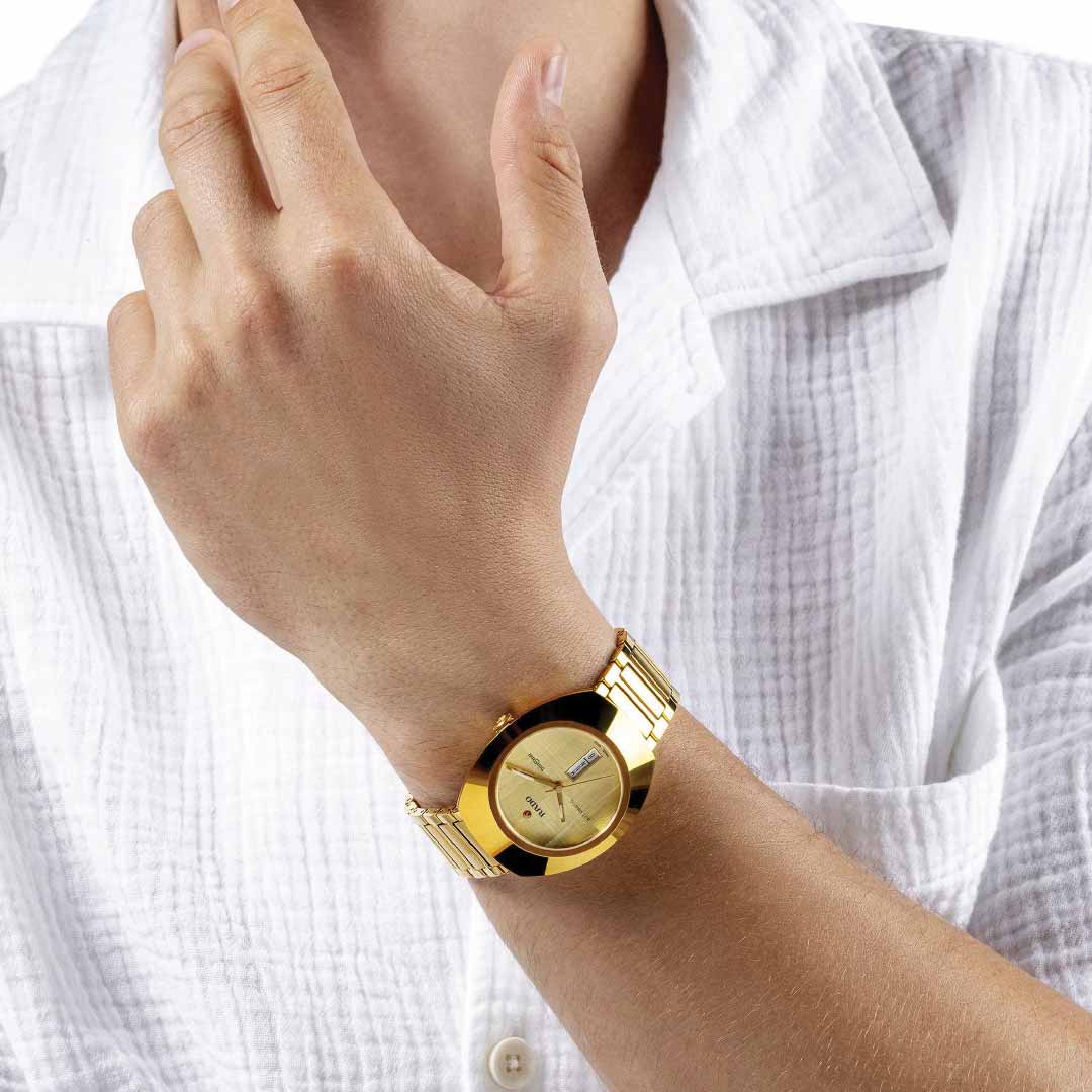 A person in a white shirt is wearing the stunning RADO DiaStar Original Automatic 38mm Watch, featuring an elegant yellow gold-coloured Ceramos™ metallic band. The watch face is beautifully complemented by silver hour and minute hands, automatic movement, and a date display. They adjust the watch with their other hand.