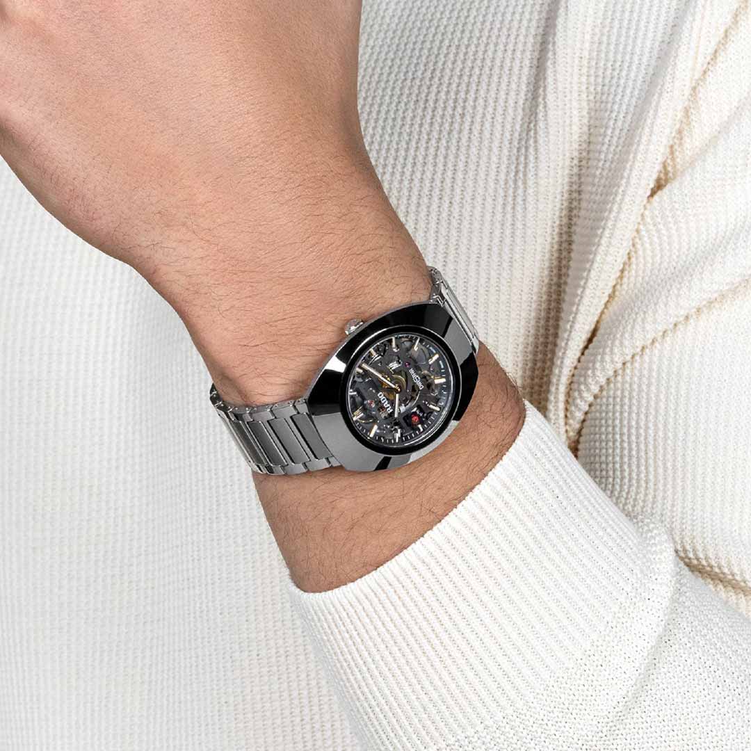 A person wearing a RADO DiaStar Original Skeleton Automatic 38mm Watch on their wrist, featuring a silver metallic design with a sapphire crystal and intricate dial, paired with a white textured long-sleeve shirt.