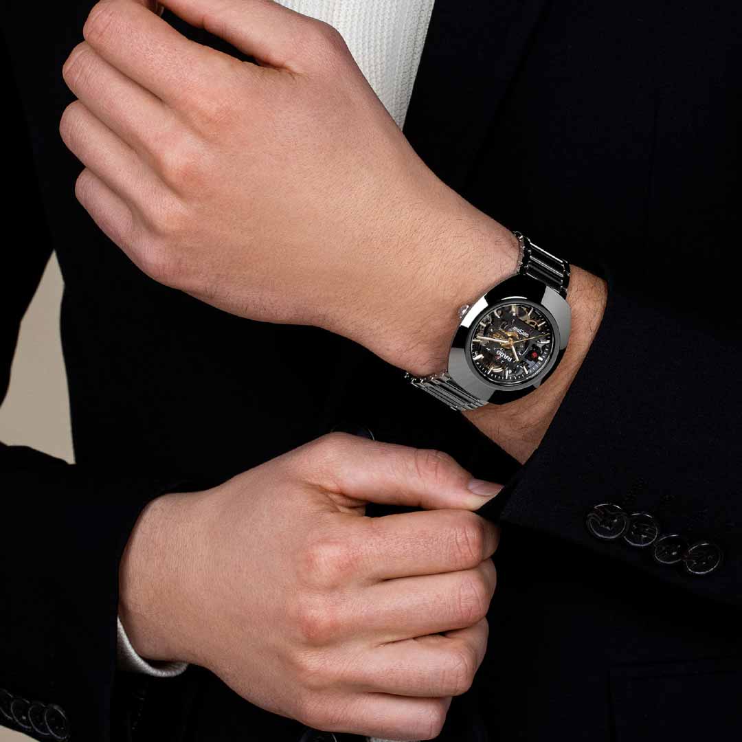 A person in a black suit is adjusting their silver wristwatch, which showcases an intricate exposed mechanical design with the R808 automatic calibre. A glimpse of a white shirt emerges from under the suit, while the neutral backdrop accentuates the elegance of this RADO DiaStar Original Skeleton Automatic 38mm Watch.