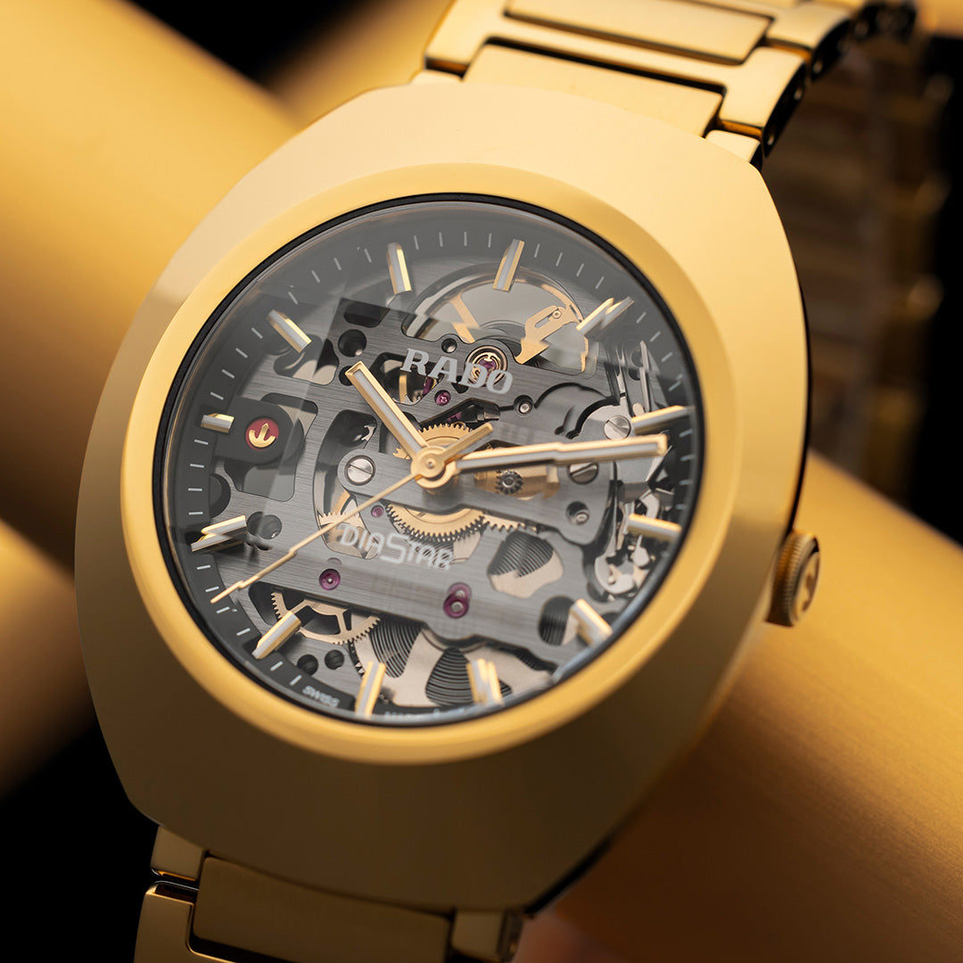 A close-up of the RADO DiaStar Original Skeleton Automatic 38mm Watch with visible gears and a gold Ceramos bezel, set against a blurred golden background.