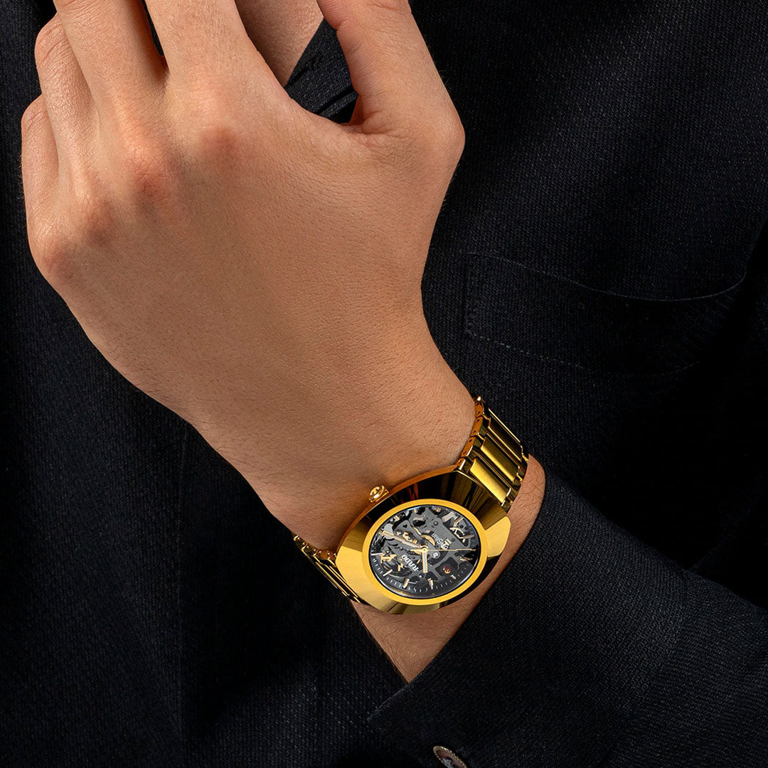 A person in a black shirt wears a RADO DiaStar Original Automatic 38mm Watch with a gold case and metal band on their left wrist, hand raised near their face, showing its visible automatic movement.