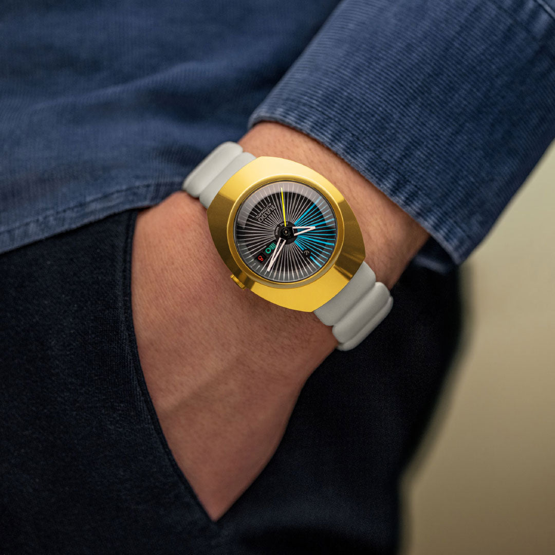 A person in blue pants and a blue shirt wears a RADO Diastar Original X Tej Chauhan Special Edition Automatic 38mm Watch with Swiss craftsmanship, a white band, and a colorful intricate dial, as their hand rests in their pocket.