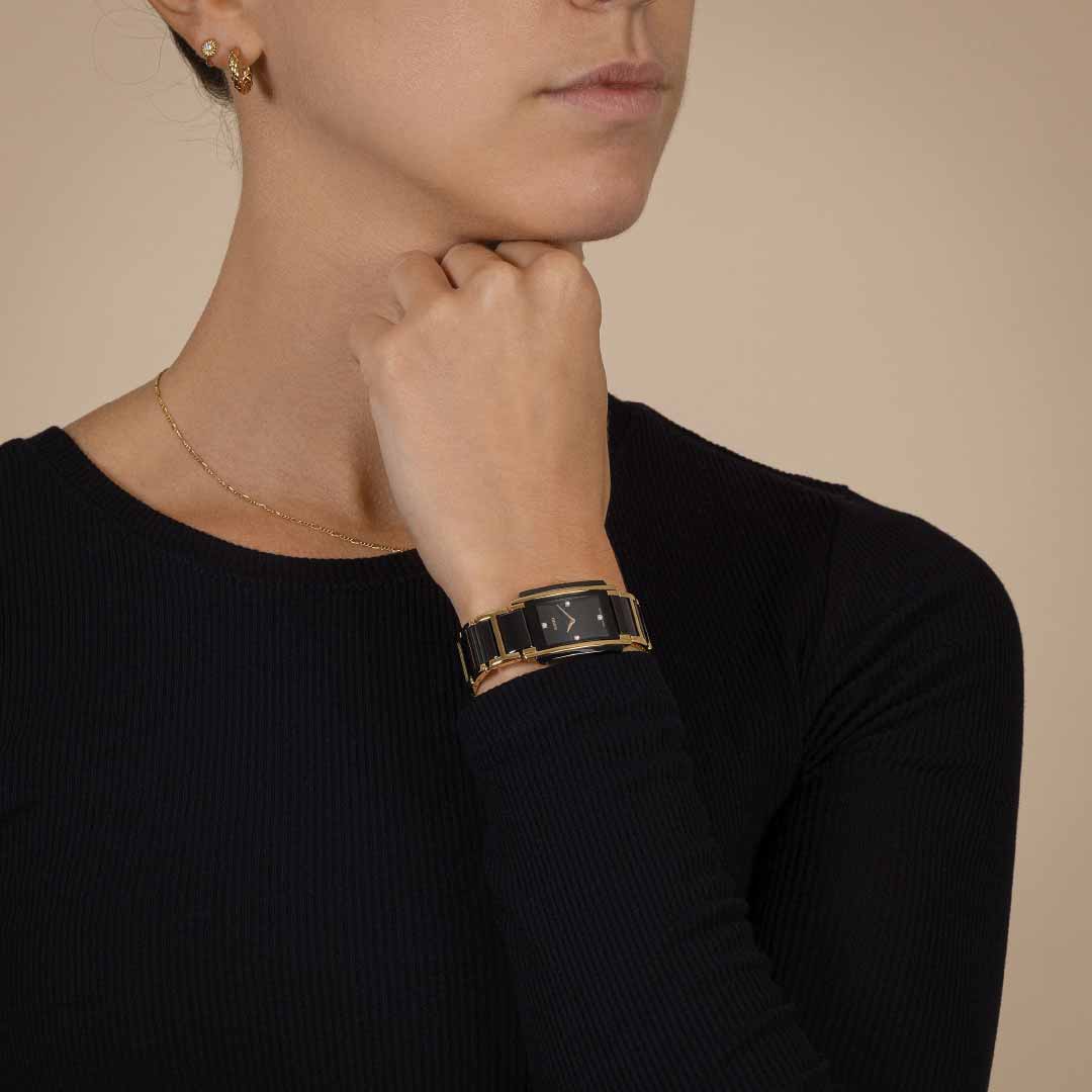 A person with short hair is dressed in a simple black outfit and is touching their chin. They accessorize with a gold chain necklace and earrings, highlighted by the RADO Integral Diamonds Quartz 22.7mm watch in black and gold ceramic on their wrist. The background features a plain light beige color.