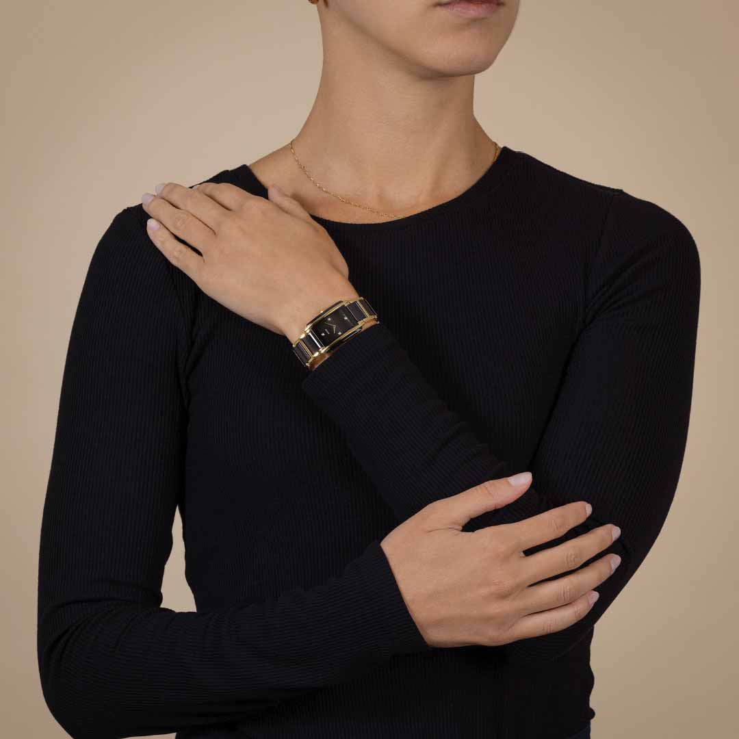 A person dressed in a black long-sleeve top is depicted with their arms crossed over their chest. On their right wrist, they wear the RADO Integral Diamonds Quartz 22.7mm Watch, featuring high-tech ceramic accents and adding a touch of sophistication. The background is plain and neutral in color.