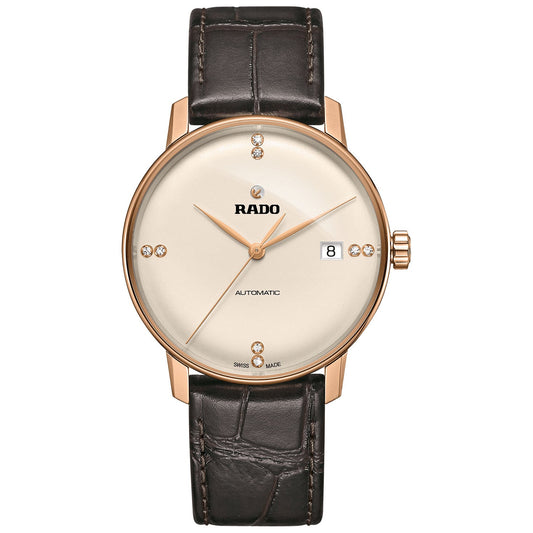 The RADO Coupole Classic Automatic 38mm Watch emanates sophistication with its gold-toned case and cream dial, prominently featuring the brand name "RADO" alongside a date display. Enhanced by sapphire crystal and diamond-like markers at key positions, it boasts an automatic movement and is complemented by a luxurious brown leather strap.
