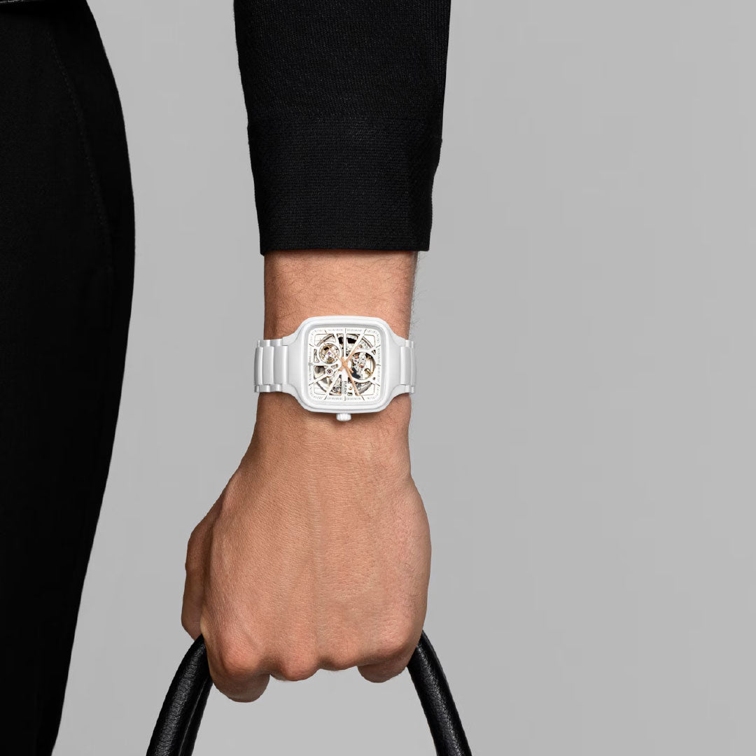 A person in black long sleeves and pants holds a black handle, displaying the RADO True Square Automatic Open Heart 38mm Watch with a white square skeleton dial on their wrist against a plain gray background.