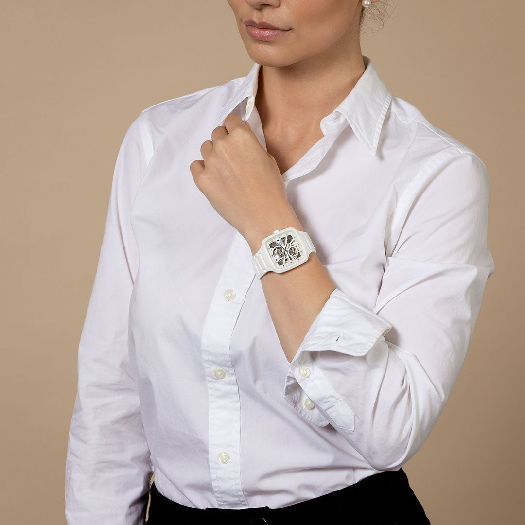 A woman in a white button-up shirt and black pants poses with her left arm raised, showcasing the RADO True Square Automatic Open Heart 38mm Watch by RADO against a plain beige background.