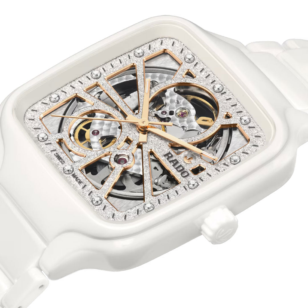A close-up of the RADO True Square Automatic Open Heart Sparkling 38mm Watch, featuring a white ceramic case, square skeletonized dial with gold accents, silver textured background, visible automatic movement, and gemstone hour markers.