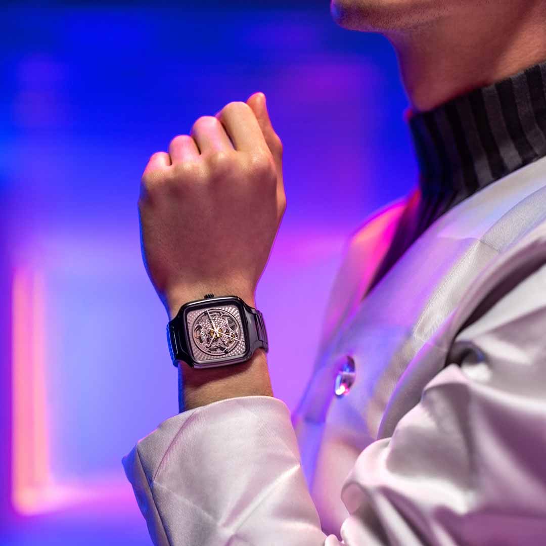 A person wearing the RADO True Square X Kunihiko Morinaga Special Edition Automatic 38mm Watch, featuring a sophisticated and sleek design with an intricate automatic calibre. The background is softly illuminated in purple and pink tones, enhancing the stylish and shiny high-tech ceramic case of the watch and complementing the person's high-collared jacket.